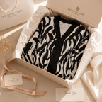 A black and white abstract patterned cardigan or wrap jacket presented in an open branded box with tissue paper, alongside matching External Links Trading packaging, ribbon, tag, and thank-you card on a warm beige surface.
