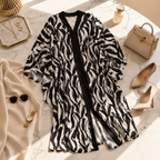 A black and white zebra-print kimono cardigan with a solid black front placket, styled in a luxury flat-lay on a white marble surface alongside beige shoes, a handbag, sunglasses, and gold accessories.