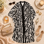 A black and white zebra-print open-front cardigan or cover-up is styled in a flat lay with fashion accessories including a beige quilted handbag, sunglasses, perfume, jewelry, sandals, and a candle on a cream fabric background.