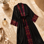 A black kimono-style robe with red, purple, and gold geometric embroidered trim along the front opening, cuffs, and tie belt, laid flat on a beige marble surface with decorative props.
