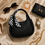 A black pebbled leather crescent-shaped handbag with a black-and-white checkered scarf tied to its handle, styled in a luxury flat lay on a beige marble surface with sunglasses, a Chanel perfume bottle, a black wallet, a gold watch, rings, a chain, and a book.