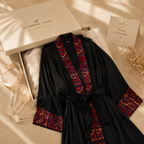A black robe with red and orange geometric embroidered trim, styled in a flat lay with an External Links gift box, ribbon, and card on a beige fabric background.
