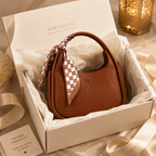 A brown pebbled leather handbag with a checkered brown-and-white scarf tied to its handle, presented inside an open white gift box with tissue paper. The box has gold text reading 'A GIFT FOR YOU' and 'Timeless Elegance, One For All.