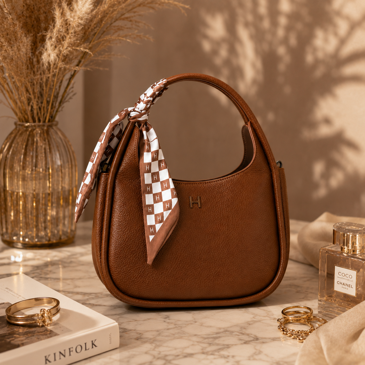 A brown pebbled leather hobo handbag with a top handle, decorated with a brown and white checkered silk scarf tied around the handle. The bag features a small gold 'H' logo on the front.