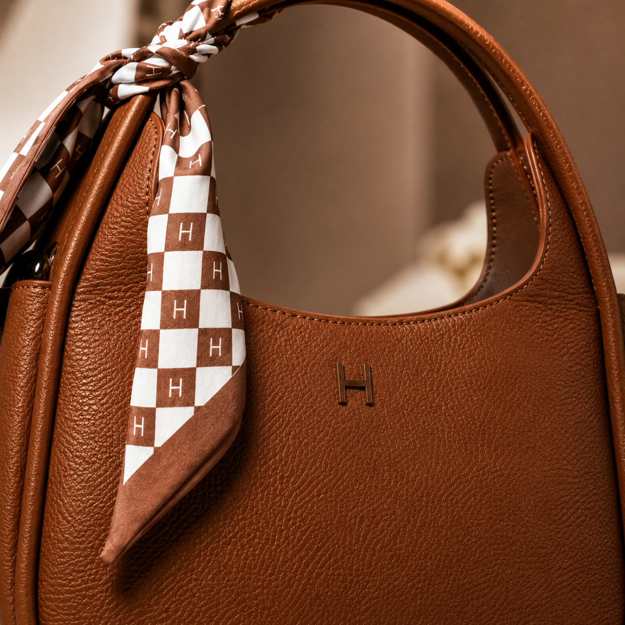 A close-up of a brown red leather handbag with a matching brown leather top handle and a tied scarf featuring a brown and white checkerboard pattern with repeating 'H' monograms. A small embossed 'H' logo is visible on the front of the bag.