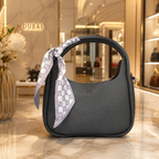 A dark gray pebbled leather handbag with a top handle, decorated with a light gray and white patterned silk scarf tied around the handle. The bag is set on a polished marble counter inside a luxury boutique with a 'DUBAI' sign in the background.