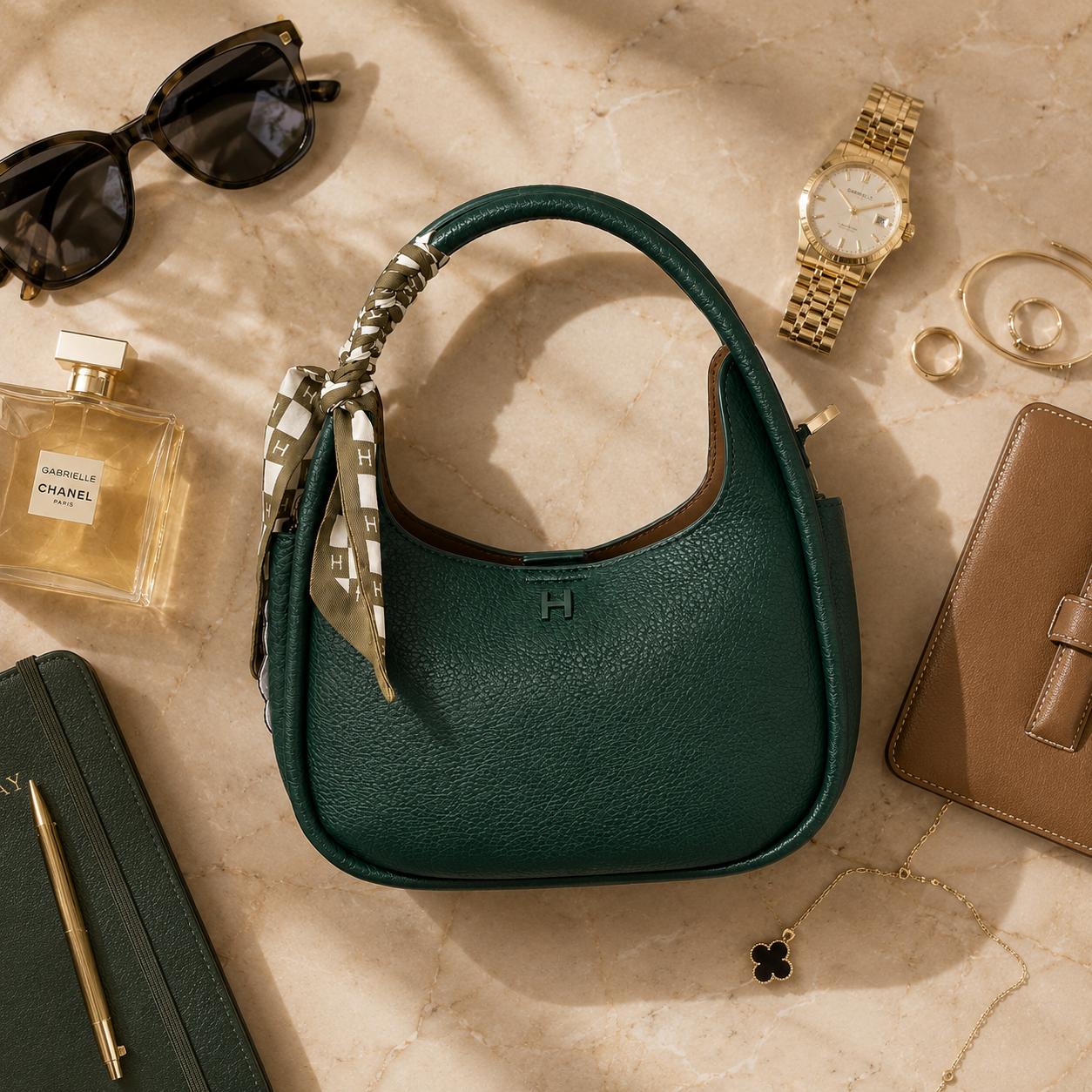 A dark green pebbled leather shoulder bag with a silver-tone handle and a patterned scarf detail, styled in a luxury flat-lay on a beige marble background with sunglasses, a perfume bottle, a gold watch, rings, and other accessories.