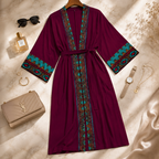 A deep burgundy kimono-style robe with teal, brown, and gold geometric patterned trim and a matching waist tie, styled in a flat lay on a beige marble surface with sunglasses, perfume, jewelry, and a handbag.