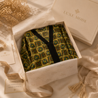 A luxury robe with a green and yellow geometric pattern and black trim, presented inside a cream-colored box with branded tissue paper and ribbon.