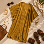 A mustard yellow kimono-style cover-up dress with a V-neck and wide draped sleeves, styled in a flat lay with sunglasses, a perfume bottle, jewelry, a watch, sandals, a handbag, and a wallet on a sunlit cream surface.
