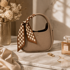 A taupe pebbled leather handbag with a matching patterned scarf tied to the handle, styled on a marble surface with dried flowers, rings, and a perfume bottle in a warm, sunlit setting.
