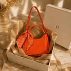 An orange crescent-shaped handbag with a matching orange monogram scarf tied in a bow, presented inside a white gift box lined with brown tissue paper. A white branded box and a thank-you card are placed beside it on a marble surface.
