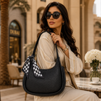A woman in a beige outfit and sunglasses carries a black pebbled leather shoulder bag with a black-and-white checkered scarf accent, set in an elegant outdoor cafe environment.