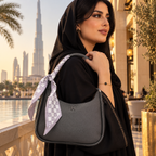 A woman in a black abaya and headscarf carries a dark gray pebbled leather shoulder bag with a patterned white and gray scarf tied to the strap, set against a Dubai cityscape background with the Burj Khalifa visible.
