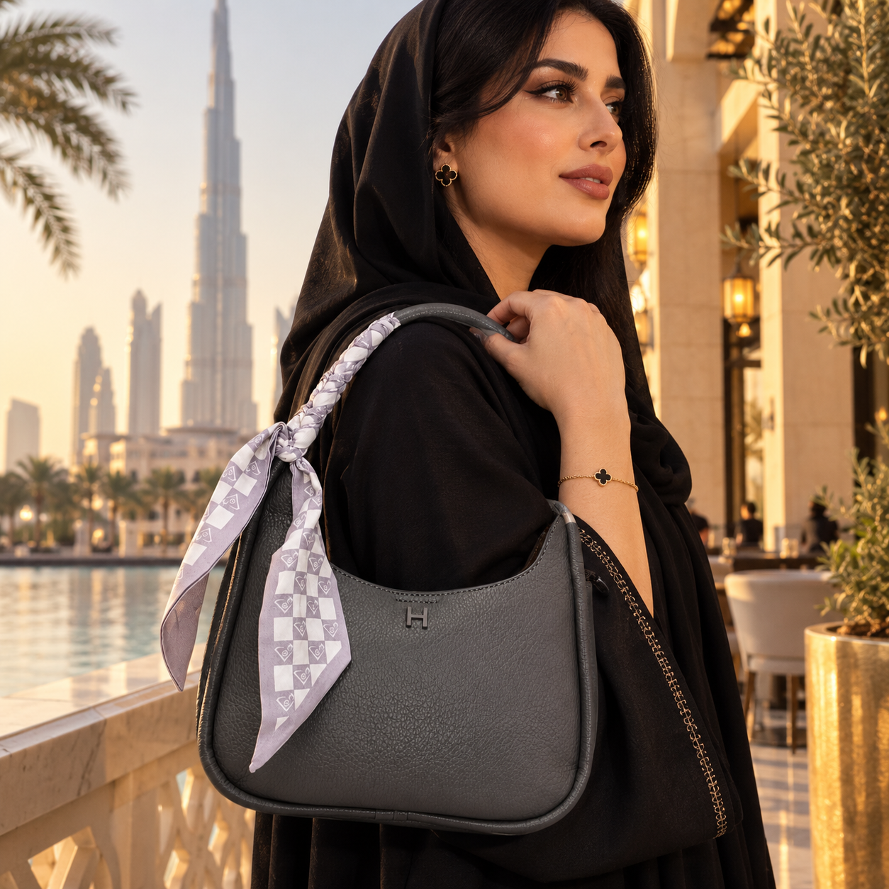 A woman in a black abaya and headscarf carries a dark gray pebbled leather shoulder bag with a patterned white and gray scarf tied to the strap, set against a Dubai cityscape background with the Burj Khalifa visible.