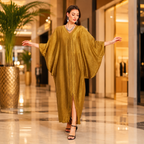 A woman modeling a flowing, loose-fitting kaftan dress in a warm mustard yellow or ochre color, walking through a luxurious indoor hallway with palm trees and warm lighting.