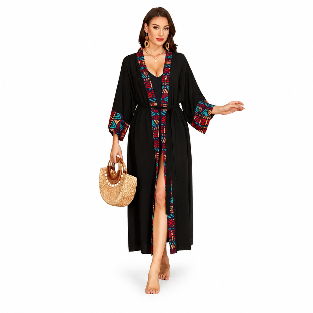 A woman modeling a long black kimono-style robe with colorful geometric trim along the neckline, front opening, and sleeve cuffs. She is barefoot and holding a woven straw handbag against a plain white studio background.