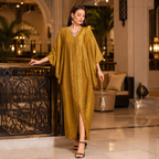 A woman models a flowing mustard yellow kaftan dress in a luxurious hotel lobby. The dress features a V-neckline, wide draped sleeves, and a high front slit.