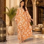 A woman models a long-sleeved maxi dress with a bold orange and white wavy abstract pattern in an elegant, sunlit indoor courtyard setting.