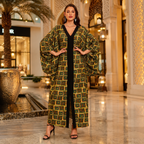 A woman models a long, wide-sleeved kaftan dress with a green, yellow, and black geometric pattern over a black V-neck dress, standing in a luxurious indoor mall setting.