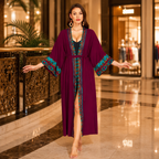 A woman stands barefoot in a luxury shopping mall corridor wearing a long, deep purple kaftan robe with teal and gold patterned trim on the sleeves, collar, and front opening.