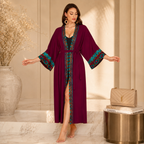 A woman stands barefoot in a warm beige interior wearing a long magenta robe with teal and multicolor embroidered trim on the sleeves and front opening. A quilted beige handbag rests on stacked books beside a round metallic side table.