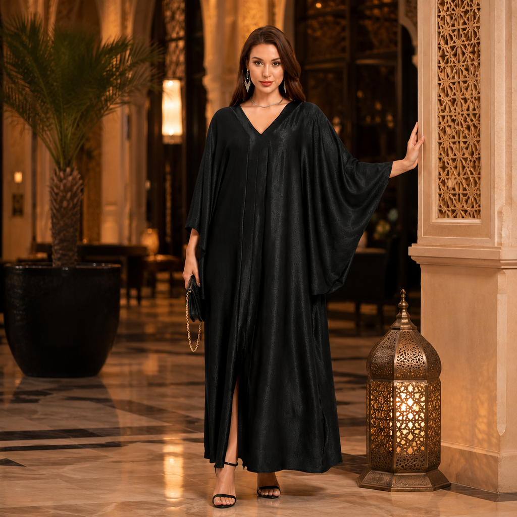 A woman stands in a grand indoor hallway wearing a flowing black caftan with wide draped sleeves and a V-neckline. The setting features warm lighting, a decorative lantern, and a potted palm in the background.