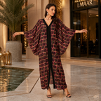 A woman stands in a luxury indoor setting wearing a long, flowy kaftan-style cover-up with a geometric pattern in dark red, black, and brown over a black jumpsuit. The background features a marble floor, a decorative pool, and a storefront with the brand name EMAAR visible.