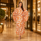 A woman stands in a luxury shopping mall corridor wearing a long, flowing caftan-style dress with a bold orange and white abstract pattern. The background features a Dubai store entrance and polished marble floors.