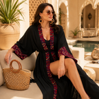 A woman wearing a black long kaftan-style cover-up with red and blue geometric patterned trim sits on a beige outdoor sofa beside a woven straw handbag, with a pool and arched architectural background.