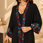A woman wearing a black open-front robe-style dress with colorful geometric patterned trim and wide patterned sleeves, styled with gold hoop earrings and a necklace in a warm indoor setting.