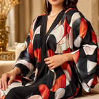 A woman wearing a loose, open-front kaftan-style robe with a bold abstract print in black, red, white, and soft pink, seated indoors against a warm beige background.