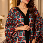 A woman wearing  a loose, open-front kaftan-style top with a bold geometric pattern in red, teal, blue, yellow, and pink over a solid black V-neck inner layer, photographed in a softly lit indoor setting.