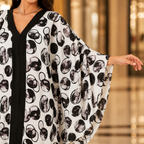 A woman wearing a white kaftan-style tunic with a black and gray abstract circular print, featuring a black V-neckline and wide flowing sleeves, photographed in a softly blurred indoor setting.
