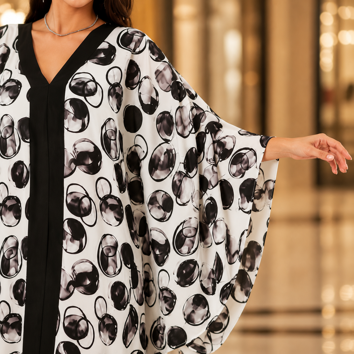 A woman wearing a white kaftan-style tunic with a black and gray abstract circular print, featuring a black V-neckline and wide flowing sleeves, photographed in a softly blurred indoor setting.