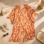 An orange and white wavy patterned short-sleeve dress styled in a flat lay on a beige marble surface, surrounded by a cream handbag, white strappy sandals, sunglasses, and gold jewelry.