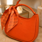 An orange textured leather shoulder bag with a matching orange silk scarf tied to the handle, set on a beige marble surface.
