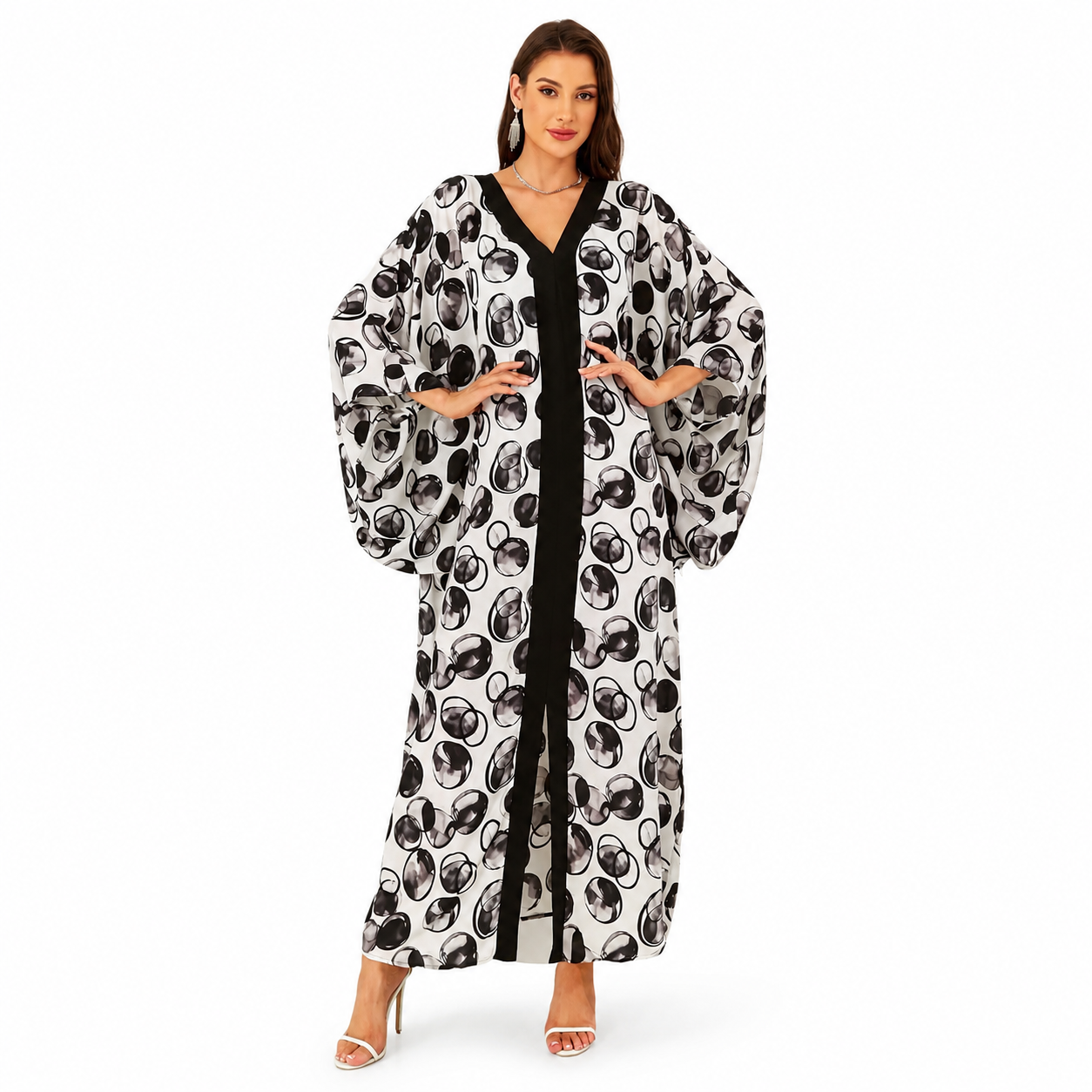 A woman modeling a long, flowing kaftan-style dress with a black and white abstract circular print over a black inner dress, photographed against a plain white studio background.