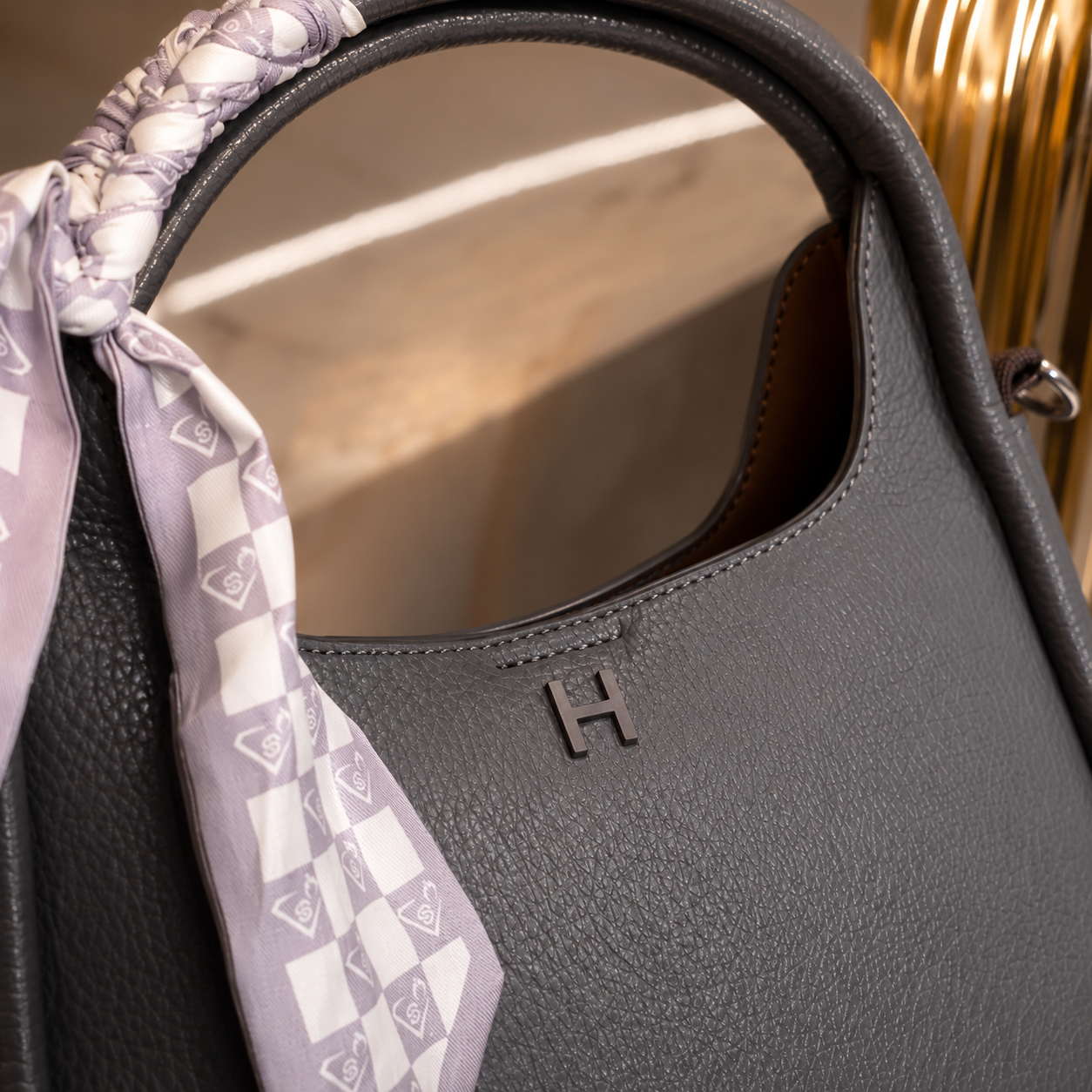 Close-up of a dark gray pebbled leather handbag with a small embossed 'H' logo, featuring a lavender and white patterned silk scarf tied to the handle.