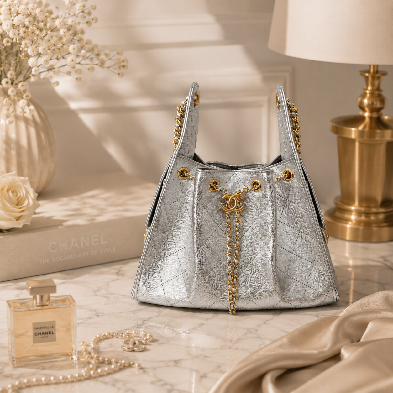 A silver quilted Chanel handbag with gold-tone chain straps and hardware, styled on a marble surface with a Chanel perfume bottle, pearl necklace, a rose, a book, and a lamp in a warm, elegant interior setting.