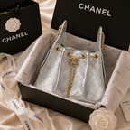 A silver quilted Chanel handbag with gold chain details, presented in an open black Chanel box with branded tissue paper, ribbon, shopping bag, and flower decorations on a satin fabric background.