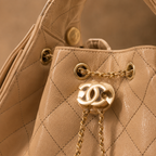 Close-up of a beige quilted leather handbag with gold-tone hardware, featuring a prominent interlocking CC logo charm and chain detail.