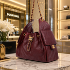 A Chanel quilted leather shoulder bag in deep burgundy, displayed on a marble counter inside a luxury boutique. The bag features gold-tone hardware, a chain strap, and a prominent interlocking CC logo charm.