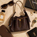 A dark brown quilted leather handbag with gold-tone chain handles and a front CC logo, styled in a luxury flat-lay with sunglasses, a Chanel perfume bottle, jewelry, a watch, a wallet, and a card holder on a light marble surface.
