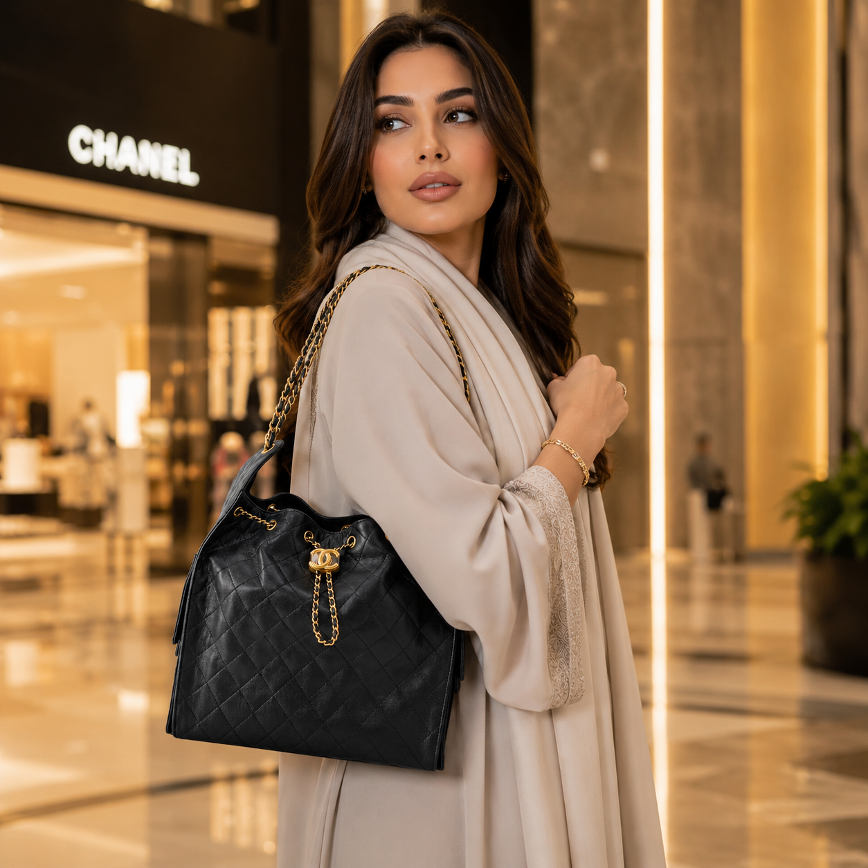 A woman in a beige coat carries a black quilted Chanel tote bag with gold hardware in a luxury shopping mall setting, with the Chanel storefront visible in the background.