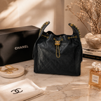 A black quilted Chanel handbag with gold-tone chain details and a gold interlocking CC charm, displayed on a marble surface with a Chanel box, dust bag, perfume bottle, and decorative props in the background.
