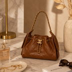 A brown quilted leather handbag with gold-tone chain straps and a prominent interlocking CC logo, styled on a marble tabletop next to a perfume bottle, sunglasses, and a vase.