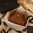 A brown quilted leather shoulder bag with gold-tone hardware and a chain strap, presented in an open black box with branded packaging, ribbons, and a box lid on a beige fabric background.