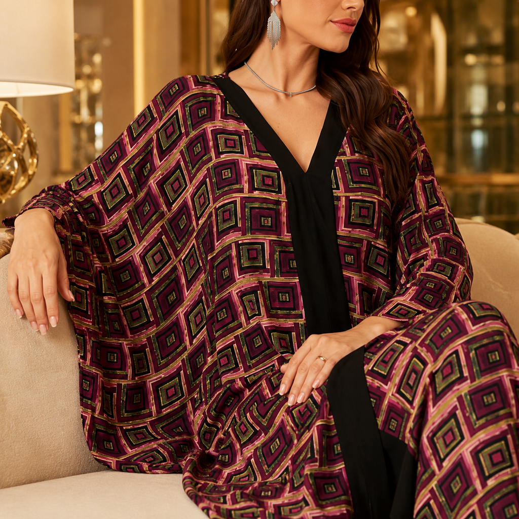 A woman seated on a beige sofa wearing a loose, draped black outfit with a bold geometric print in purple, magenta, and green. The setting appears to be an indoor living space with warm lighting and blurred background decor.