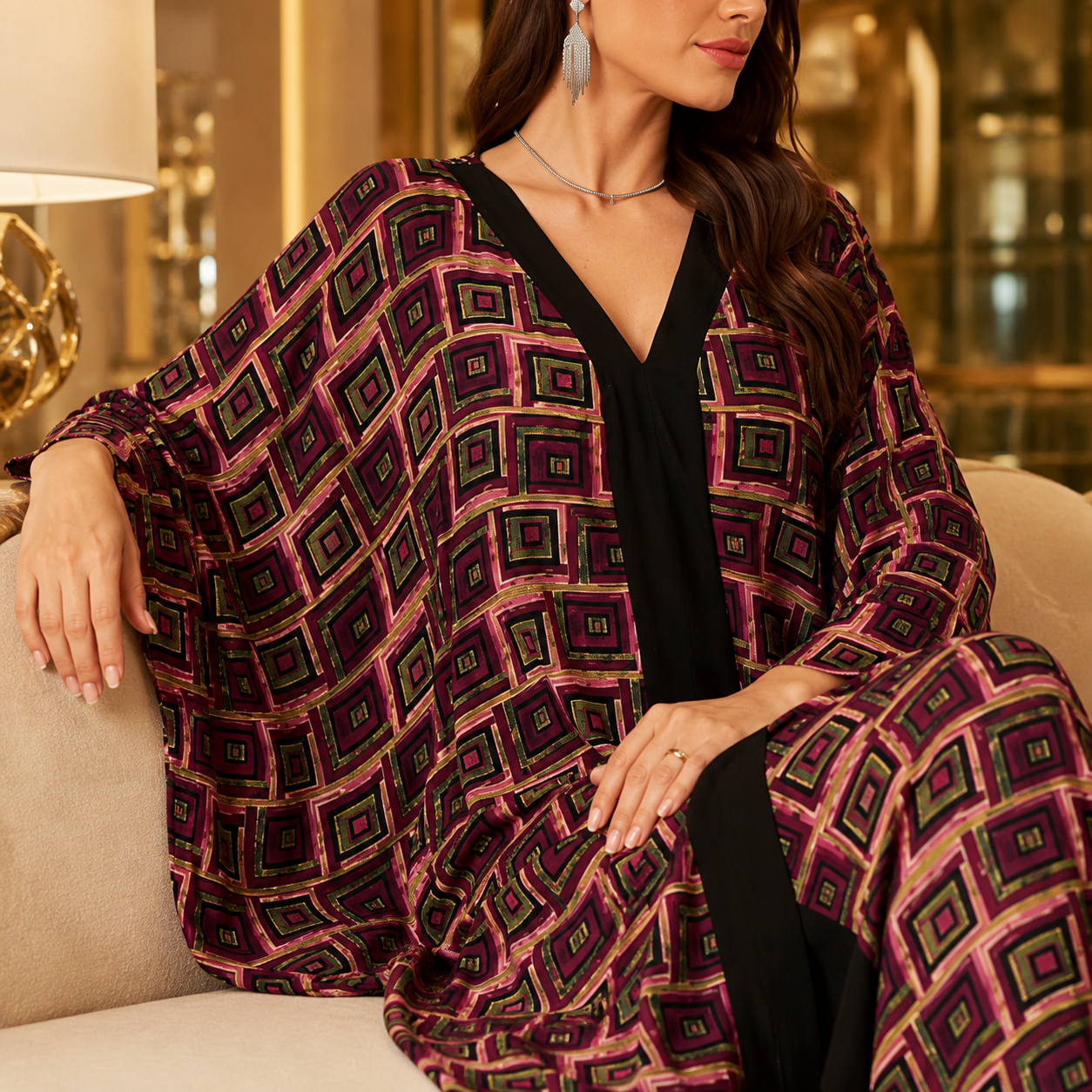 A woman seated on a beige sofa wearing a loose, draped black outfit with a bold geometric print in purple, magenta, and green. The setting appears to be an indoor living space with warm lighting and blurred background decor.